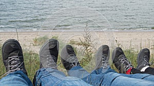 People sitting with legs outstretched on hill, three pairs of feet spread in gray sneakers on backdrop of grass and blue sea
