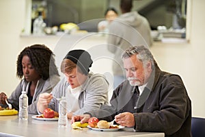 People Sitting At Table Eating Food In Homeless Shelter