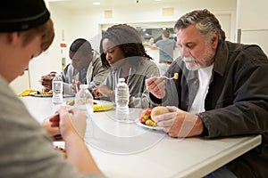 People Sitting At Table Eating Food In Homeless Shelter