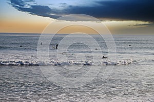 People surfing in vast blue ocean water with waves rolling into the beach with clear blue sky at sunset