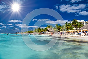People swimming near white sand beach with umbrellas, bungalow bar and cocos palms, turquoise caribbean sea, Isla Mujeres island,
