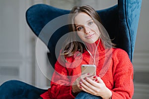 People and technology concept. Satisfied female meloman enjoys favourite music, listens audio record, dressed in red sweater, surf