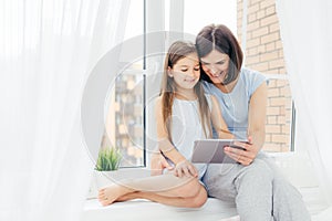 People, technology, family, children concept. Positive young other and her small daughter sit on window sill, hold digital tablet,