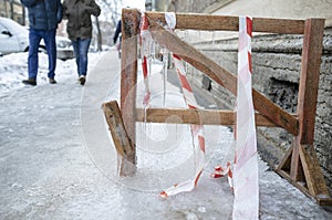 People walk on the icy traumatic sidewalk, which is frozen homemade wooden obstacle, with icicles hanging from it and a protective