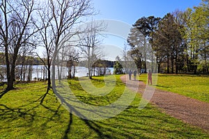 People walking down a footpath in the park near blue lake water with lush green and autumn colored trees and lush green grass