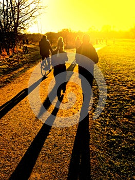 People walking on a path in a park casting long shadows in the intense and glowing bright low sunshine.
