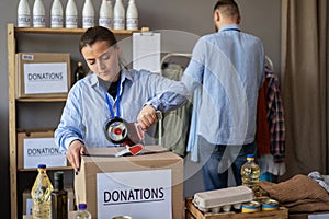 People working in charitable foundation. Female volunteer with scotch dispenser closing package near carton boxes in