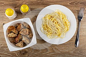 Pepper and salt, white bowl with pieces fried pork meat, plate with spaghetti, fork on table. Top view