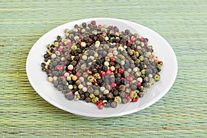 Peppercorn melange. White, black, red and green pepper grains on a white saucer on a green table mat made of natural plant fibers photo