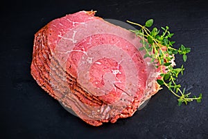 Peppered roast beef pastrami slices on black stone background with herbs