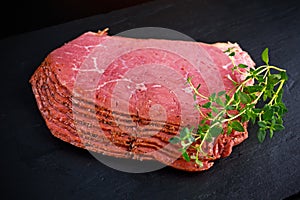 Peppered roast beef pastrami slices on black stone background with herbs
