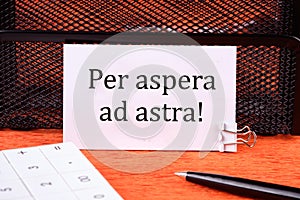 Per aspera as astra in English means through hardships to the stars on a clean white business card next to a calculator, a pen on