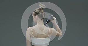 Percussion massager, young female massaging shoulder on gray studio backdrop, therapy for muscle relaxation, back view