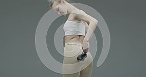 Percussion massager, young fit female massaging buttock to stimulate blood circulation on gray studio background