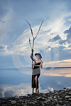 Perfect brunette beauty woman in a black hat and a black dress poses near a lake against a blue sky. Long hair woman and beautiful