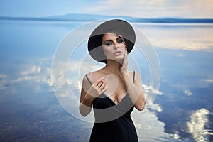 Perfect brunette beauty woman in a black hat and a black dress poses near a lake against a blue sky. Long hair woman and beautiful