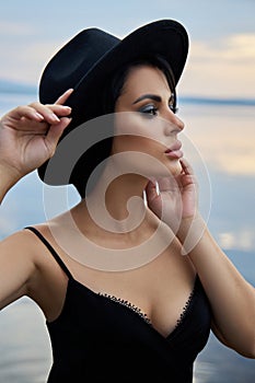 Perfect brunette beauty woman in a black hat and a black dress poses near a lake against a blue sky. Long hair woman and beautiful