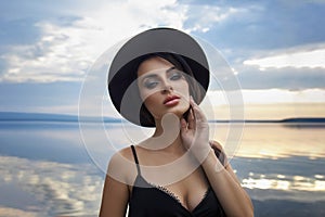 Perfect brunette beauty woman in a black hat and a black dress poses near a lake against a blue sky. Long hair woman and beautiful