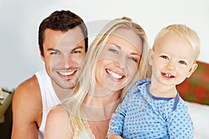 Perfect family portrait. Two parents smiiling at the camera with their young son while they sit on their bed.