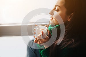 Perfect morning A young brunette woman sits on a windowsill and holds a cup of tea or coffee in her hands. Female model