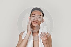 Perfect skin. Portrait of shirtless young asian man with towel around his neck looking at camera, applying lotion after
