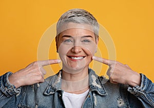 Perfect Smile. Mature woman pointing on strong healthy teeth, yellow background