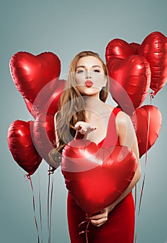 Perfect woman blowing kiss. Pretty girl with red lips makeup and red heart balloons. Fashion model wearing red dress. Love, gifts
