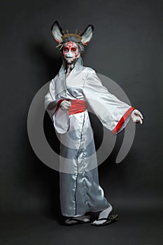 Perfect young woman wearing animal mask and white kimono standing on black background, full length portrait. Halloween, Carnival