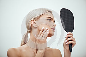 Perfection. Profile portrait of young attractive woman touching her face and looking in small mirror while standing
