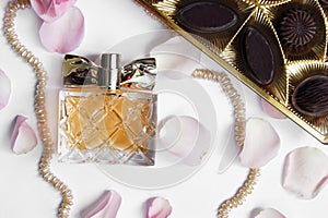 Perfume bottle with rose petals, pearls and chocolates on a light background.