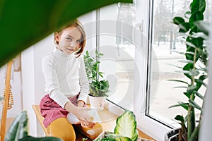 Perky looking girl sitting on a window sill, spraying house plants with water