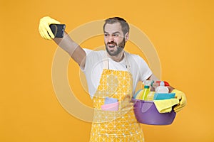 Perplexed man househusband in apron rubber gloves hold basin detergent bottles washing cleansers doing housework