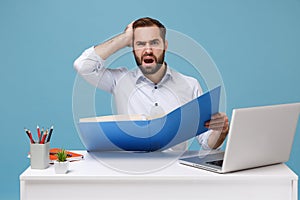 Perplexed young bearded man in light shirt sit work at desk with pc laptop isolated on pastel blue background