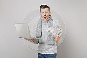 Perplexed young man in gray sweater, scarf holding lots bunch of dollars banknotes cash money, laptop pc computer