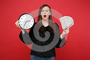 Perplexed young woman in black fur sweater holding round clock, fan of money in dollar banknotes cash money isolated on