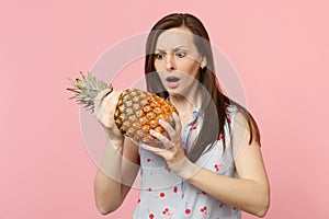 Perplexed young woman in summer clothes holding in hand fresh ripe pineapple fruit isolated on pink pastel wall