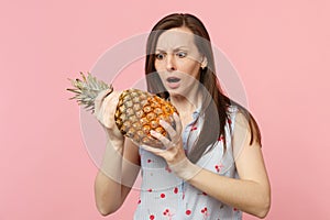 Perplexed young woman in summer clothes holding in hand fresh ripe pineapple fruit  on pink pastel wall