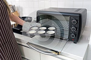 A person in an apron and oven gloves, placing a muffin tray with batter into a black toaster oven in a white kitchen