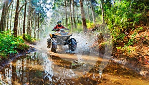 A person on an ATV is riding through a muddy trail in a forest, splashing mud