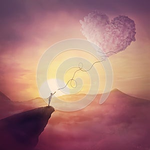 A person on the cliff and a heart shaped cloud like a kite raising up in the air. Magical scene, love and romance concept. Pink