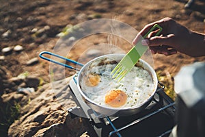 Person cooking fried eggs in nature camping outdoor, cooker prepare omelette breakfast picnic on metal gas stove, tourism recreat