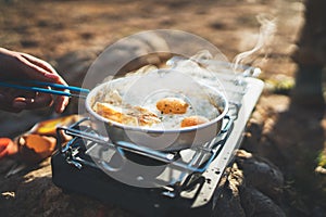 Person cooking fried eggs in nature camping outdoor, cooker prepare scrambled omelette breakfast picnic on metal stove, tourist