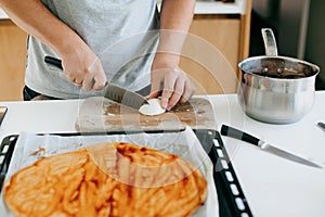 Person cutting onion on background of round dough with ketchup for pizza on modern white kitchen. Process of making home pizza,