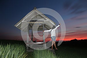 Person in the field after sunset. Happy adventure woman standing alone holding the hut in the farm adoring the sunset colors. photo
