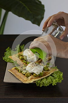 Person grinder pepper on a waffles with pouched eggs, avocado and lettuce on a wooden table background