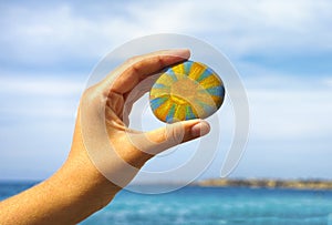 Person hand hold pebble with painted bright sun against blue sky