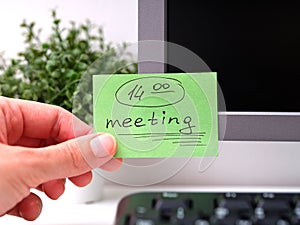 Person hand sticking a green paper note with the reminder Meeting 14-00 on it on to a monitor at an office workplace