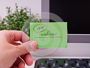 A person hand sticking a green paper note with the reminder Webinar 19-00 on it on to a monitor at office workplace