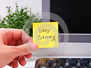 A Person hand sticking a yellow paper notes with the reminder Stay Strong on it on to a monitor at an office workplace