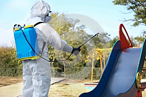 Person in hazmat suit with disinfectant sprayer near slide at children`s playground. Surface treatment during coronavirus pandemi